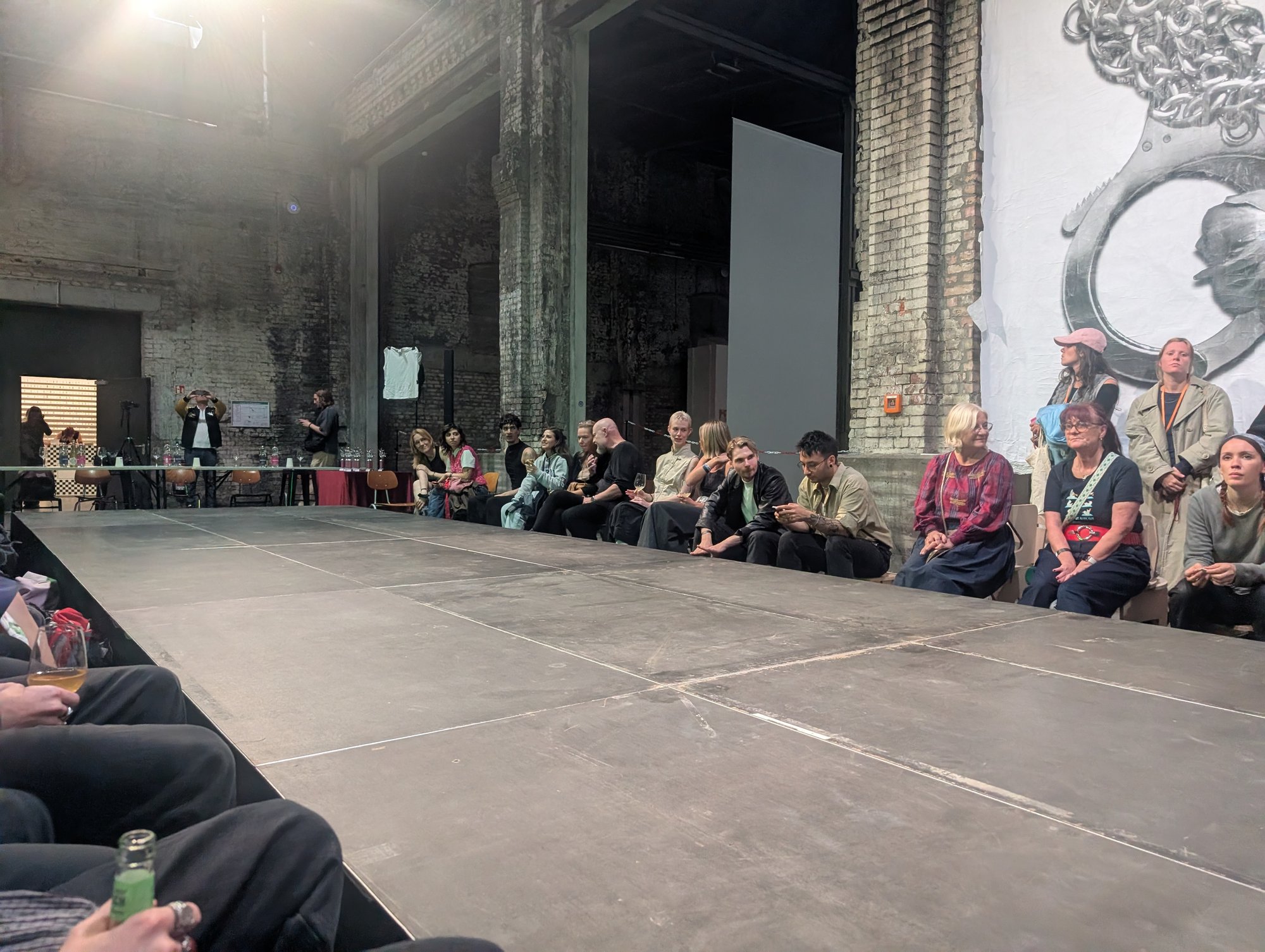 Publikum an der Kante der grauen Bühnenfläche, an der Wand ein großes Blatt mit Handschellen, HALLEN 07