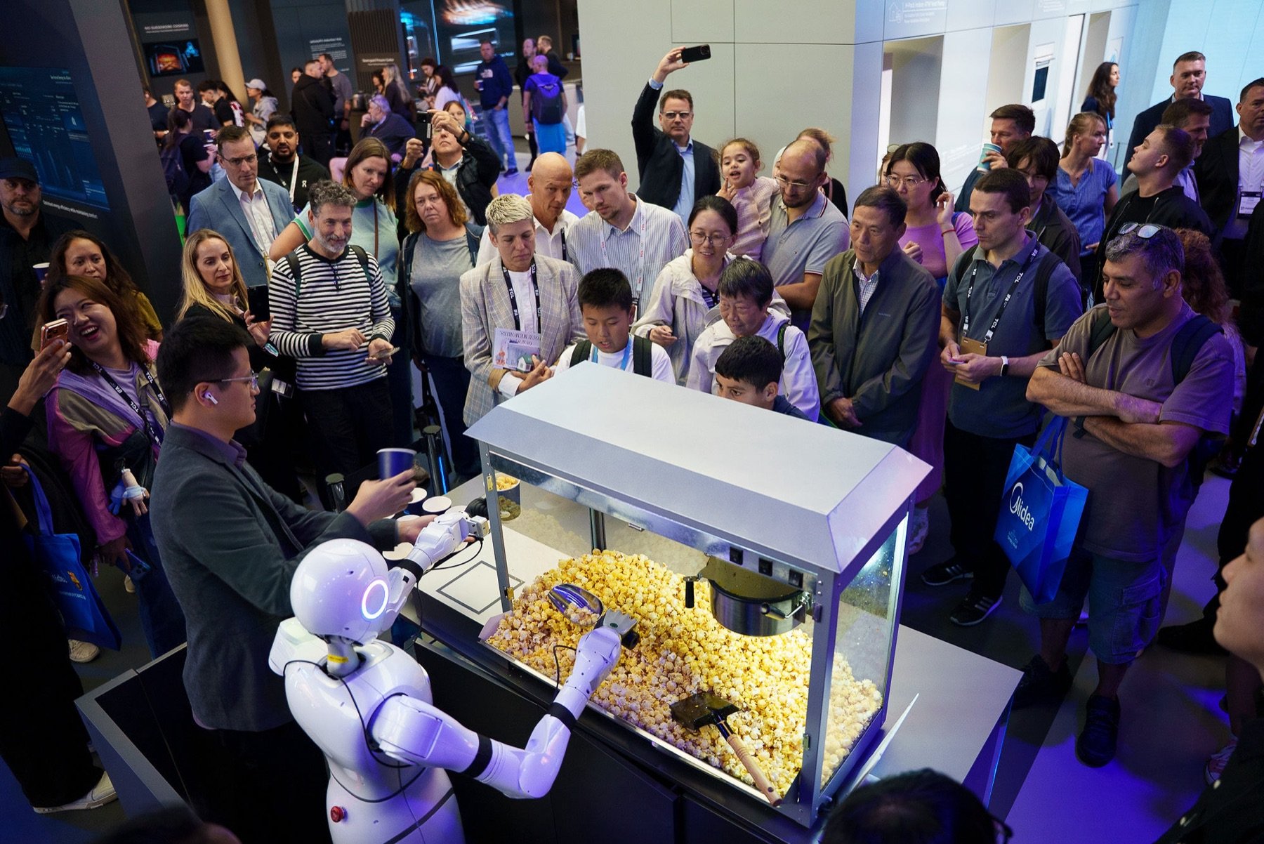 Ein weißer Humanoid schöpft Popcorn aus einer Maschine, davor Publikum mit Handys